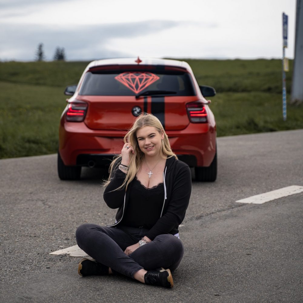 Eine blonde Frau vor einem orangenen Bmw 1er sitzend