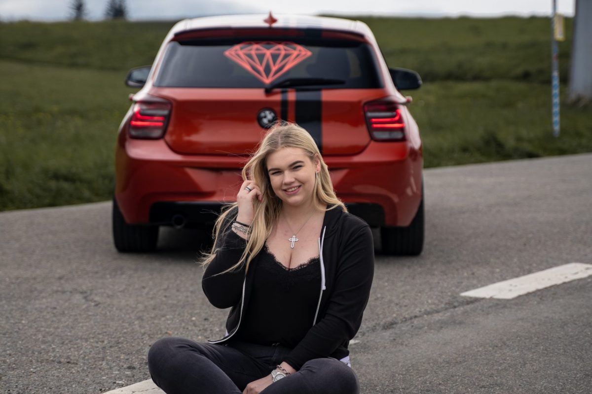 Eine blonde Frau vor einem orangenen Bmw 1er sitzend